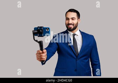 Attractive young adult bearded man blogger holding steadicam with phone, making video or has livestream, wearing official style suit. Indoor studio shot isolated on gray background. Stock Photo
