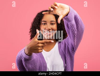 Cheerful black woman making picture frame with fingers, looking at camera and smiling for photo on pink background Stock Photo