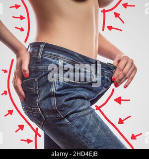 Closeup of slim waist of young woman standing in big jeans showing successful weight loss, indoor studio shot, isolated on light gray background, diet concept. Stock Photo
