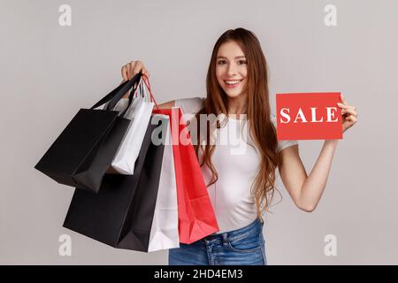 Portrait of happy beautiful smiling dark haired woman holding paper bags and sale sign, shopping and discounts, wearing white T-shirt. Indoor studio shot isolated on gray background. Stock Photo