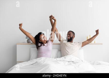 Satisfied young african american guy and lady raise hands, stretching body on bed in bedroom interior Stock Photo