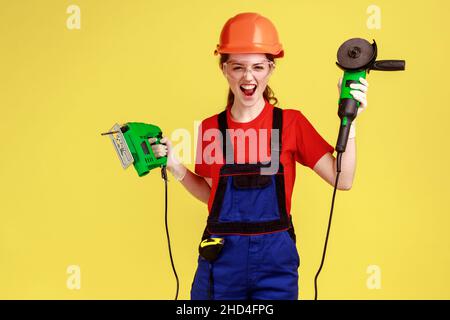 Crazy builder woman holding grinding saw and fretsaw in hands, having crazy facial expression, screaming, wearing overalls and protective helmet. Indoor studio shot isolated on yellow background. Stock Photo