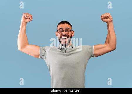 Win and triumph concept. Overjoyed arab man yelling with clenched fists, raised up in the air, blue background Stock Photo