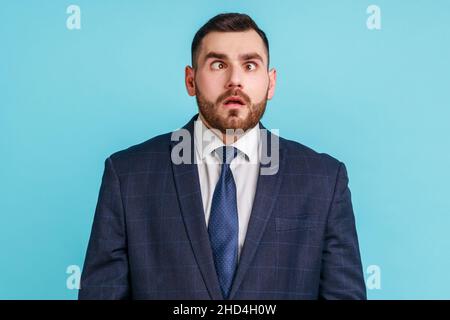 Funny amused man with beard wearing official style suit crossing his eyes looking crazy and stupid, fooling around, having fun, vision problems. Indoor studio shot isolated on blue background. Stock Photo