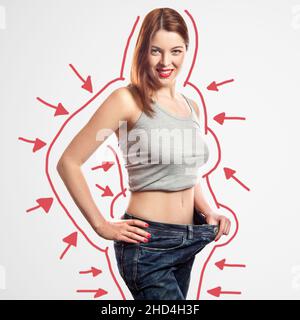 portrait of happy beautiful slim young woman in big jeans and gray top showing successful weight loss, indoor, studio shot, isolated on light gray background, diet concept. Stock Photo