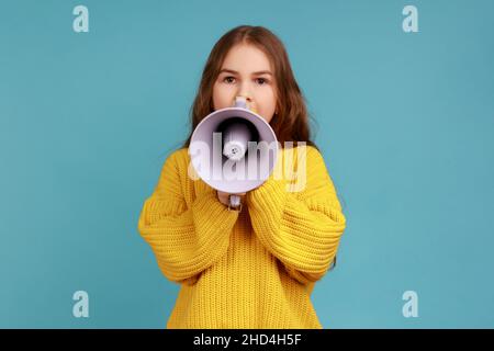 Portrait of little girl announces important school information, looks at camera with calm expression, wearing yellow casual style sweater. Indoor studio shot isolated on blue background. Stock Photo