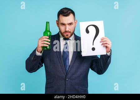 Drink or not. Portrait of serious man with beard wearing dark official style suit holding bottle with alcohol and paper with question mark. Indoor studio shot isolated on blue background. Stock Photo