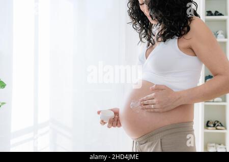 Young pretty pregnant woman with curly hair putting moisturizer on her tummy to prevent stretch marks in pregnancy.White background. copy space Stock Photo