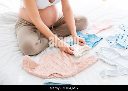 pregnant young woman sitting on bed folding and organizing clothes for her future baby Stock Photo