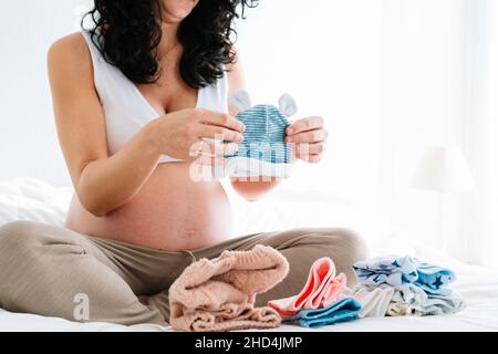 young pregnant woman with curly hair sitting on the bed looking at a little hat for her future newborn baby. folded baby clothes on the bed Stock Photo