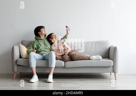 Split system. Happy young Asian couple with remote relaxing under air conditioner, sitting on couch at home, mockup Stock Photo