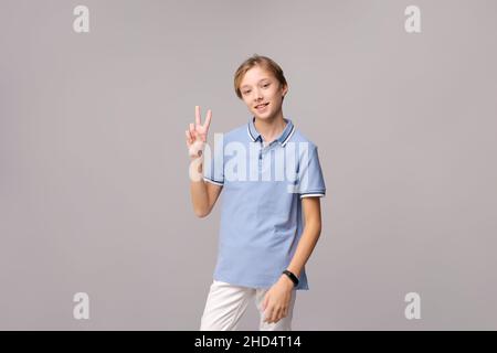 Caucasian young guy in blue shirt happy on light background shows victory gesture with fingers. Handsome smile of a teenage guy. The concept of luck in achieving the goal Stock Photo