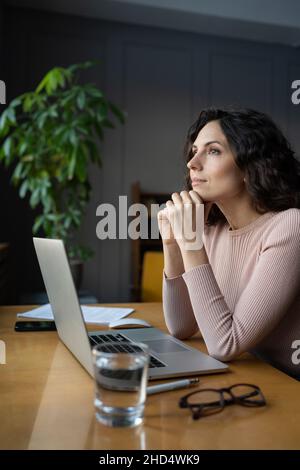 Young thoughtful italian woman employee sitting at office desk with laptop looking through window Stock Photo