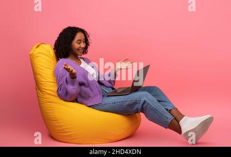 Smiling young black woman making video call with friends, using laptop, sitting in bean bag chair on pink background Stock Photo