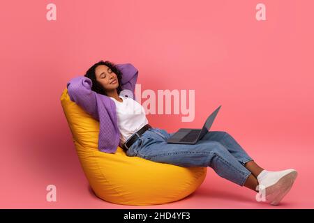 Young black woman with laptop taking break, sitting in bean bag chair with closed eyes, relaxing after working online Stock Photo