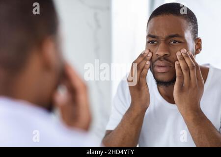 Dull Skin. Worried Young Black Man Looking At Mirror And Touching Face Stock Photo