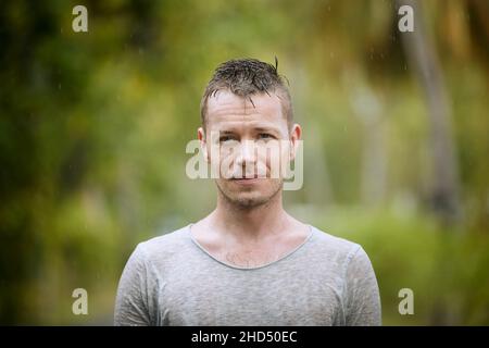 Portrait in heavy rain. Drenched young man with drops on face looking at camera. Stock Photo