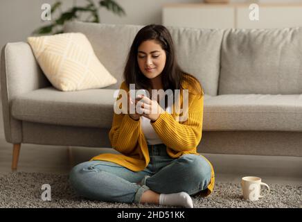 Positive young arab woman reading online morning news on smartphone and drinking coffee, sitting on floor in living room Stock Photo