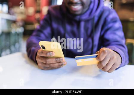 Crop positive African American male client sitting at table in cafe and using cellphone for online shopping and paying with plastic card Stock Photo