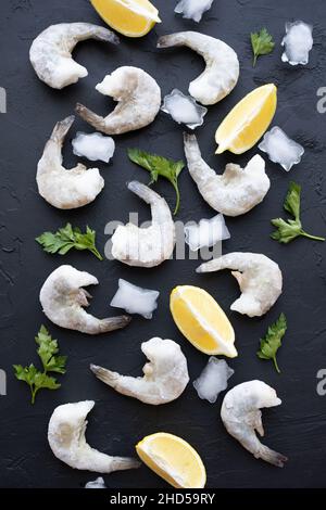 Seafood with lemon, parsley and ice on dark surface. Frozen raw shrimps. King Prawns on black stone background. Served food, preparing healthy food Stock Photo