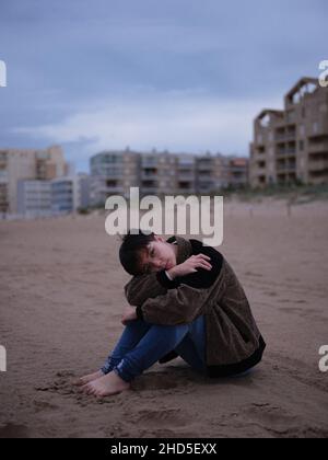 non binary young woman alone on the beach sitting on the sand Stock Photo