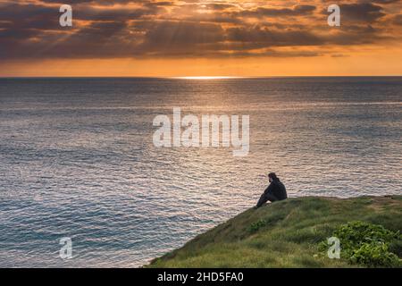 A man sitting on Towan Head enjoying a spectacular sunset over Fistral Bay in Newquay in Cornwall. Stock Photo
