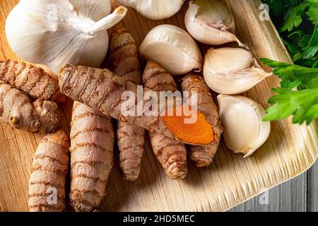 Raw organic curcuma roots and garlic cloves on a wooden plate close-up. Fresh whole turmeric rhizomes and garlic as spices, seasonings, antioxidant. Stock Photo