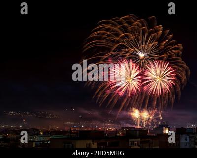 Bright New Year fireworks sparkles in the dark sky over city. The small town celebrates with colorful fireworks at night. Outdoor night scene. Winter. Stock Photo