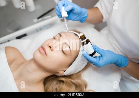 Hands of cosmetologist dripping nourishing oil facial skin of young woman in spa salon. Facial treatment and skincare concept Stock Photo