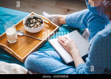 Woman writes in large white open notebook, sitting on bed in blue pajamas with breakfast with oatmeal. Top view. Morning routine. Healthy lifestyle Stock Photo