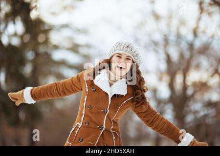 smiling modern middle aged woman with mittens in a knitted hat and sheepskin coat rejoicing outdoors in the city park in winter. Stock Photo