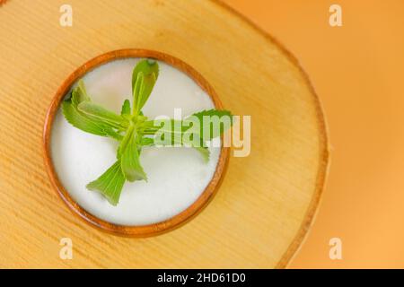 Stevia fresh green twig in a wooden round cup with crystal stevia powder on a wooden saw cut on a orange background. Stock Photo