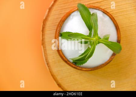 Stevia rebaudiana on orange background.Stevia twig in a wooden round cup with crystal stevia powder on a wooden saw cut on a orange background. Stock Photo