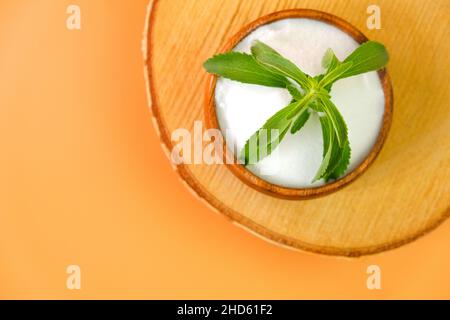 Stevia rebaudiana on orange background.Stevia fresh green twig in a wooden cup with crystal stevia powder on a wooden saw cut Stock Photo