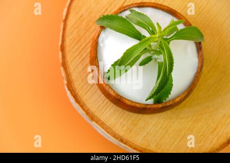 Stevia rebaudiana on orange background.Stevia fresh green twig in a wooden round cup with crystal stevia powder on a wooden saw cut on a orange Stock Photo