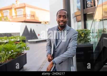 Smiling black businessman is elegantly dressed and is holding his laptop. Confident and happy business manager. Established millennial person Stock Photo