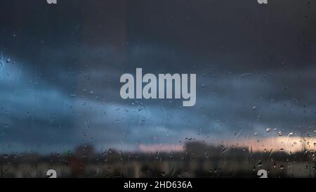 Rainy day through the window on cloudy grey sky and city buildings background. Evening cityscape behind the glass window with trickling drops of water Stock Photo