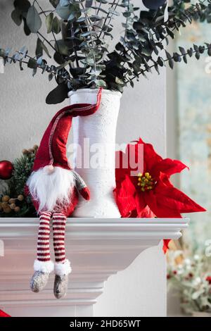 A classic norweigan holiday Christmas gnome decoration sitting on a white modern mantle with a white vase with eucalyptus and a poinsettia bloom Stock Photo