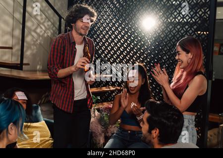 Cheerful and handsome male friend standing holding beer bottle with sticky note on forehead with group of people clapping and laughing at home during party Stock Photo