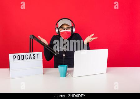 Presenter man wearing red cap and mask in podcast radio recording studio, next to a computer, a microphone and a light box with the word podcast, upse Stock Photo