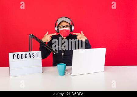Presenter man wearing red cap and mask in podcast radio recording studio, next to a computer, a microphone and a light box with the word podcast, poin Stock Photo