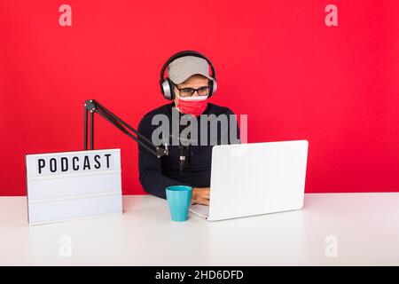Presenter man wearing red cap and mask in podcast radio recording studio, next to a computer, a microphone and a light box with the word podcast, cons Stock Photo