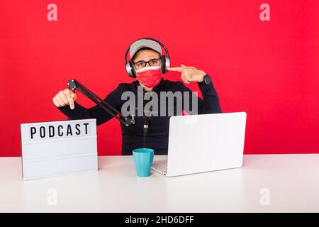 Presenter man wearing red cap and mask in podcast radio recording studio, next to a computer, a microphone and a light box with the word podcast, poin Stock Photo