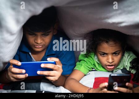 Kids busy playing video game on mobile phone at night under the bedsheet - concept smartphone and technology addiction, unhealthy childhood lifestyle Stock Photo