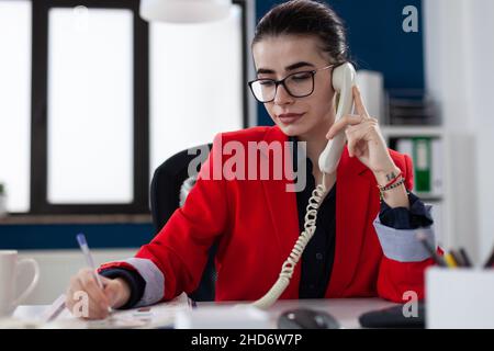 Entrepreneur taking notes in phonecall sitting at desk. Businesswoman with glasses working in startup office. Employee in red jacket writing on paper in telephone conversation. Stock Photo