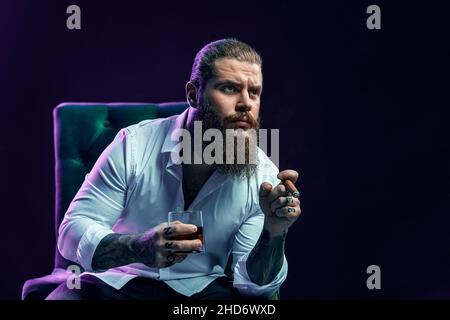 Portrait of thinking bearded man who smoke cigar holds glass of whiskey while sittingin armchair.  Stock Photo