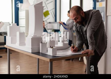 Development engineer making notes on construction plan of residential project leaning over desk with maquette. Architect drawing blueprints in architectural office next to white foam scale model. Stock Photo