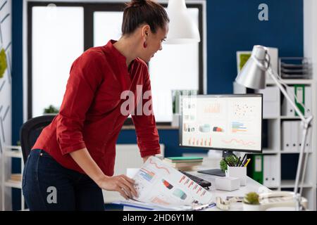 Entrepreneur leaning over desk looking at charts on desktop computer screen. Businesswoman standing comparing business data with clipboard. Employee in red shirt looking at pc monitor. Stock Photo