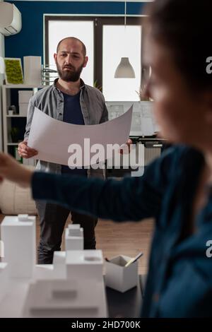 Standing architect holding blueprints thinking about architectural design working together with colleague engineer in modern office. Architects doing creative teamwork on 3d buildings maquette. Stock Photo