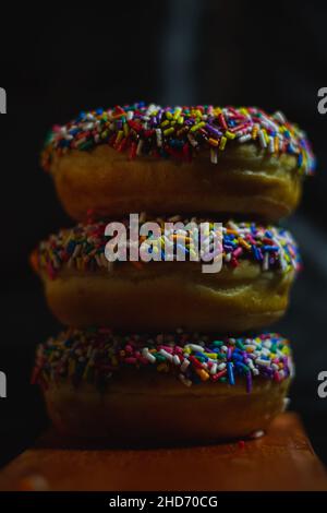 Vertical shot of three delicious donuts with sprinkles on top of each other on a wooden board Stock Photo